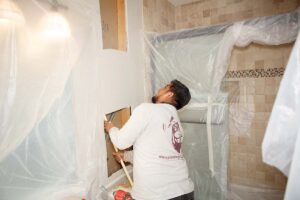 Plumber feeding pipe between two sheetrock openings in bathroom with protective plastic on walls