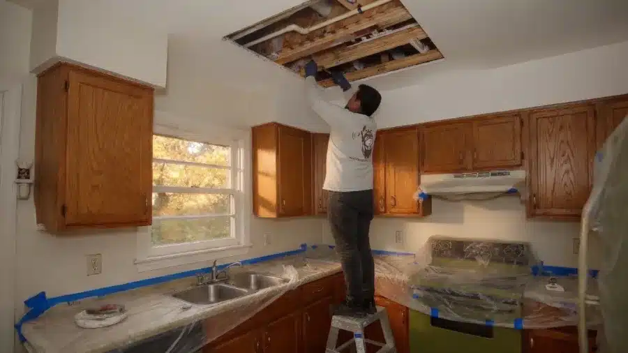 A Plumbing Express technician running new repipe lines through an opened kitchen ceiling, with counters and appliances covered in plastic.